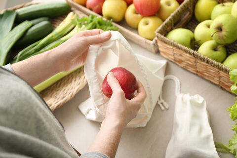 Nachhaltiger Konsum: Eine Frau Steht an einem Tisch auch dem frischen Obst und Gemüse liegt. Sie hat einen Apfel in der Hand den Sie in einen Stoffbeutel packt.